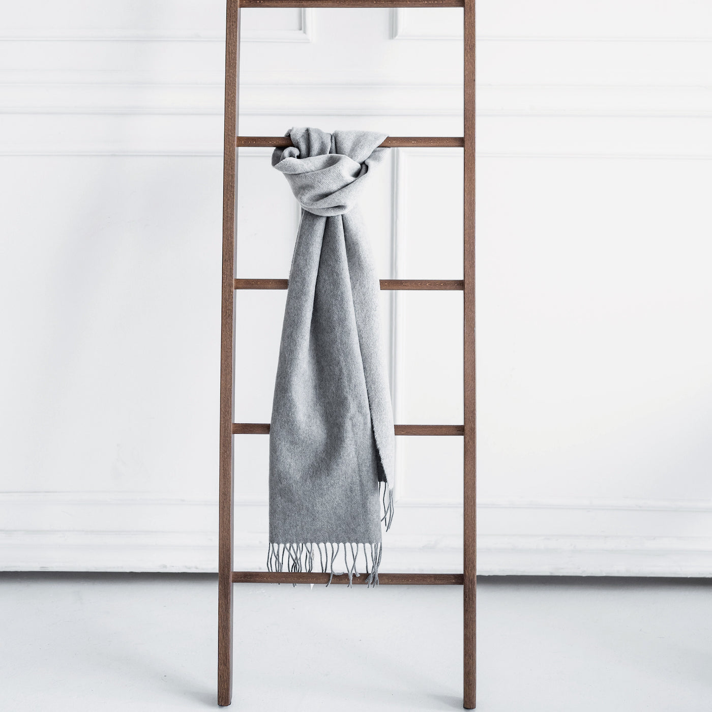 Produktbild mit grauem Kaschmir Schal and brauner Holzleiter angebunden. Im Hintergrund ist eine weiße Stuckwand zu sehen.